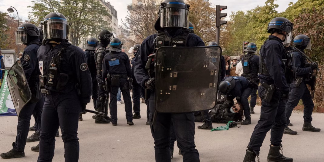 French riot police in helmets and shields standing in a line on a Paris street during a protest.